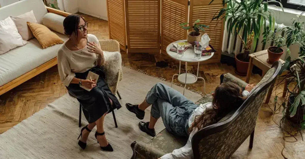 Two women engaged in a psychotherapy session in a warm, inviting interior with plants and natural lighting.