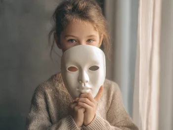 Fille autiste tenant un masque blanc devant son visage, regard sérieux, illustrant le camouflage des difficultés et des émotions vécues à l’école, dans une atmosphère calme et introspective. Filles autistes