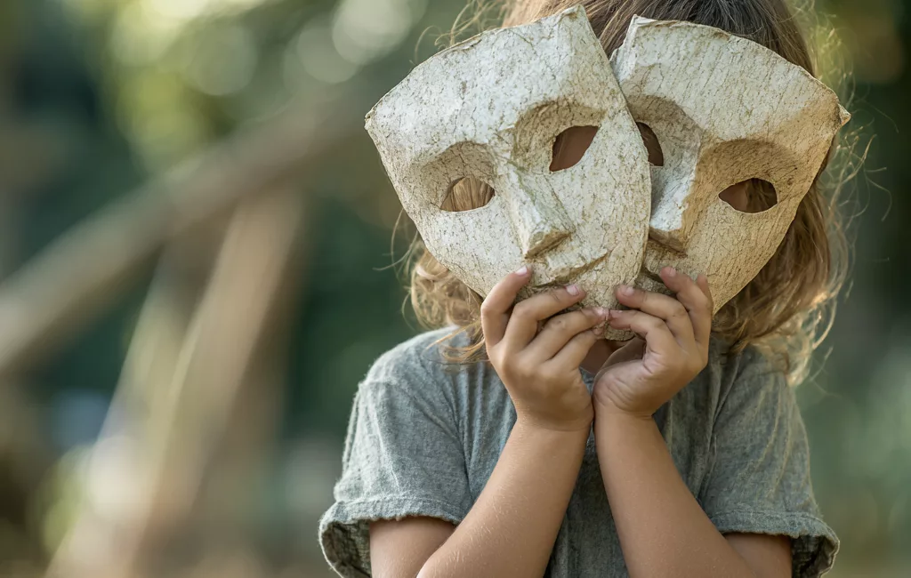 Enfant dans un parc tenant des masques de théâtre devant son visage, illustrant le masquage et l’adaptation sociale.