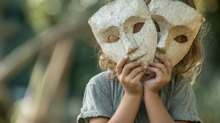 Enfant dans un parc tenant des masques de théâtre devant son visage, illustrant le masquage et l’adaptation sociale.
