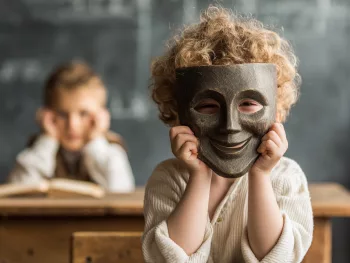 Enfant assis à son bureau à l’école portant un masque souriant, illustrant le camouflage autistique et le contraste entre l’apparence attendue en classe et le vécu intérieur.