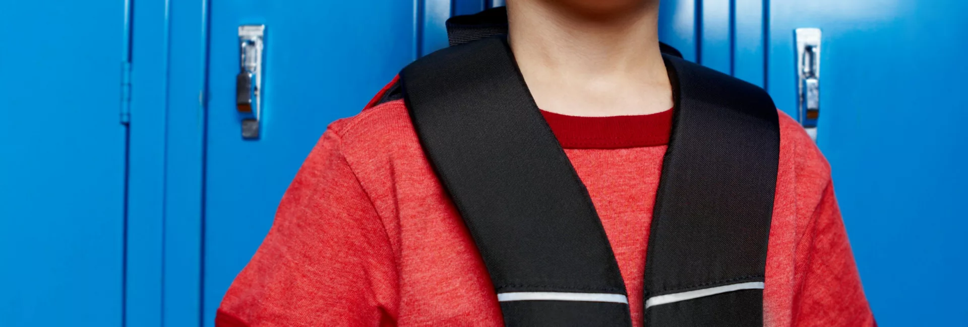 Un jeune garçon aux cheveux bruns, en tee-shirt rouge, tient les bretelles de son sac à dos noir et sourit légèrement, adossé à une rangée de casiers d'école bleu vif.