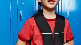 Un jeune garçon aux cheveux bruns, en tee-shirt rouge, tient les bretelles de son sac à dos noir et sourit légèrement, adossé à une rangée de casiers d'école bleu vif.