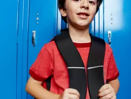 Un jeune garçon aux cheveux bruns, en tee-shirt rouge, tient les bretelles de son sac à dos noir et sourit légèrement, adossé à une rangée de casiers d'école bleu vif.