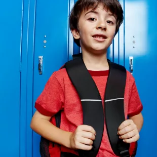 Un jeune garçon aux cheveux bruns, en tee-shirt rouge, tient les bretelles de son sac à dos noir et sourit légèrement, adossé à une rangée de casiers d'école bleu vif.