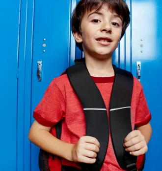 Un jeune garçon aux cheveux bruns, en tee-shirt rouge, tient les bretelles de son sac à dos noir et sourit légèrement, adossé à une rangée de casiers d'école bleu vif.