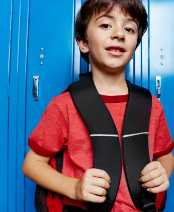 Un jeune garçon aux cheveux bruns, en tee-shirt rouge, tient les bretelles de son sac à dos noir et sourit légèrement, adossé à une rangée de casiers d'école bleu vif.