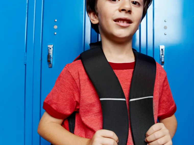 Un jeune garçon aux cheveux bruns, en tee-shirt rouge, tient les bretelles de son sac à dos noir et sourit légèrement, adossé à une rangée de casiers d'école bleu vif.