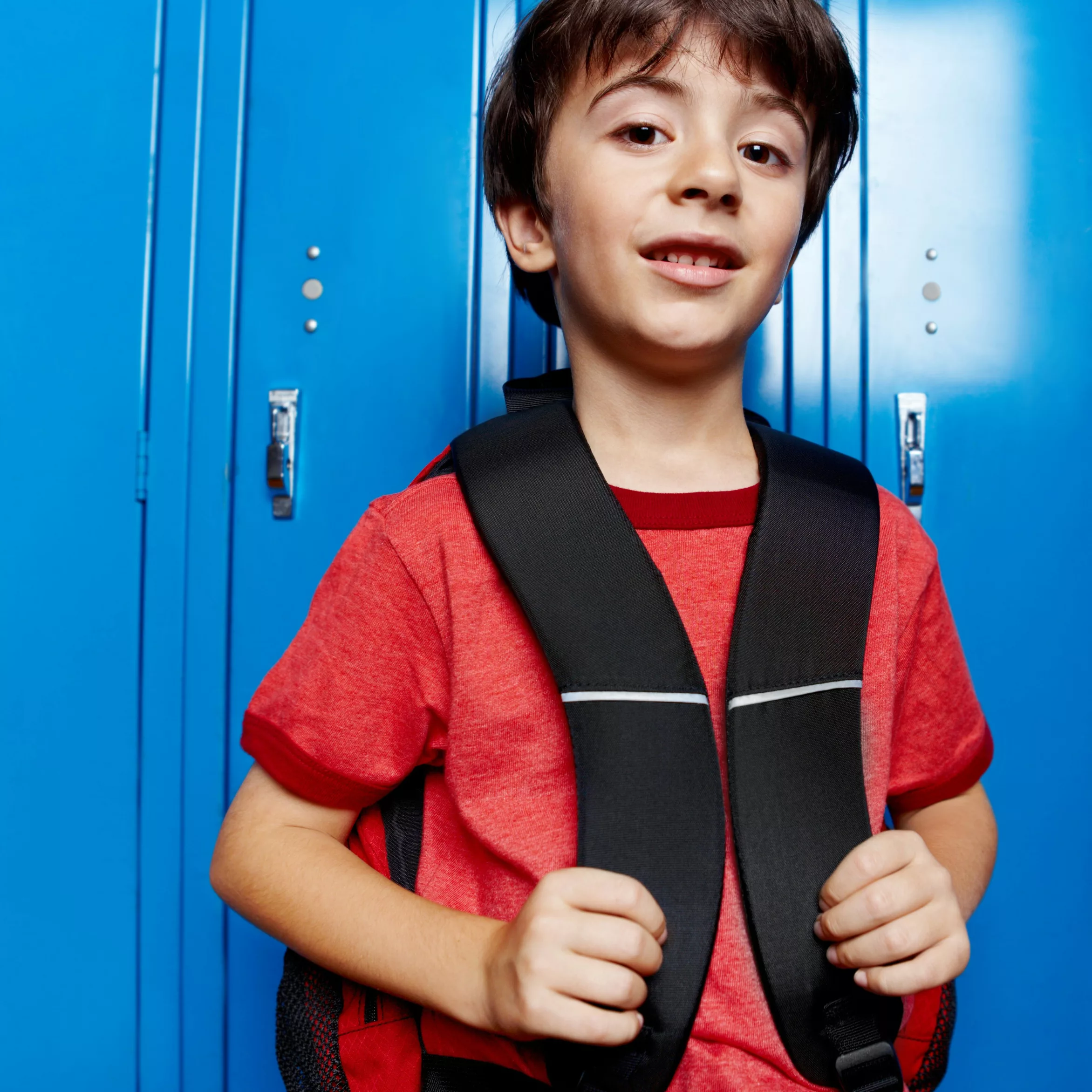 Un jeune garçon aux cheveux bruns, en tee-shirt rouge, tient les bretelles de son sac à dos noir et sourit légèrement, adossé à une rangée de casiers d'école bleu vif.