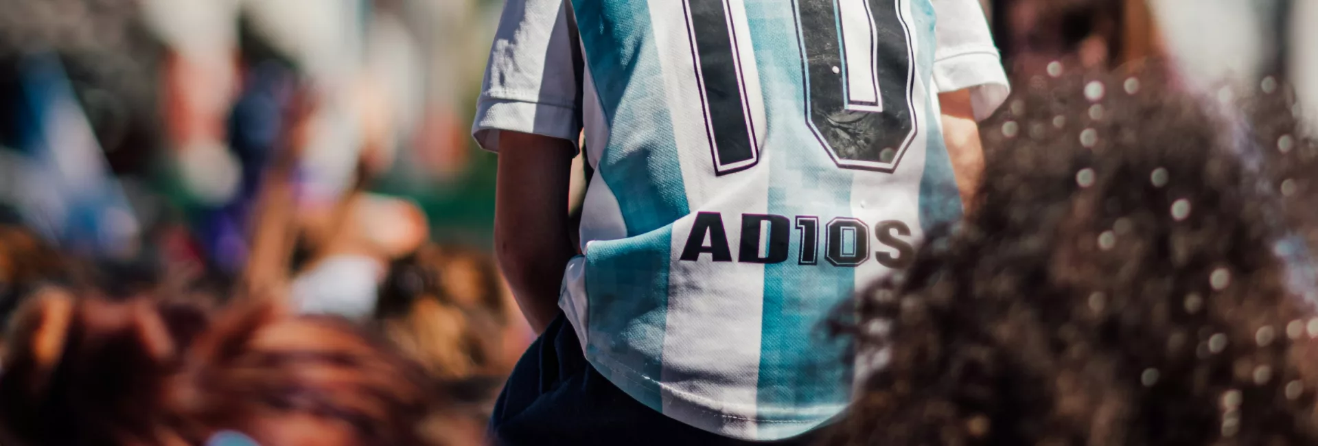 Enfant en maillot de foot argentin « Maradona 10 » sur les épaules d'un adulte, dans une foule ensoleillée.