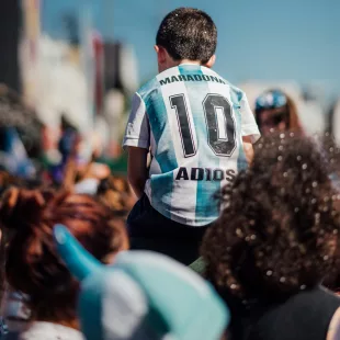 Enfant en maillot de foot argentin « Maradona 10 » sur les épaules d'un adulte, dans une foule ensoleillée.