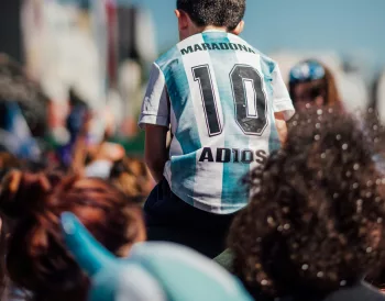 Enfant en maillot de foot argentin « Maradona 10 » sur les épaules d'un adulte, dans une foule ensoleillée.