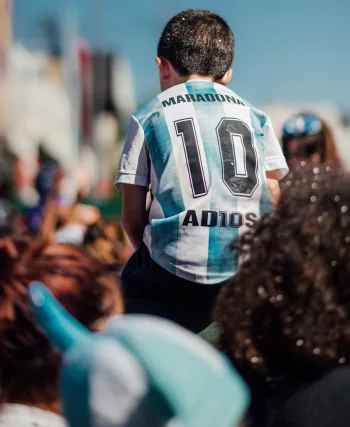 Enfant en maillot de foot argentin « Maradona 10 » sur les épaules d'un adulte, dans une foule ensoleillée.