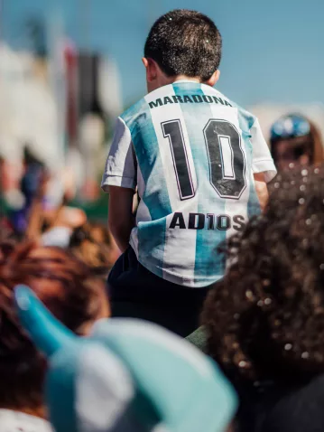 Enfant en maillot de foot argentin « Maradona 10 » sur les épaules d'un adulte, dans une foule ensoleillée.