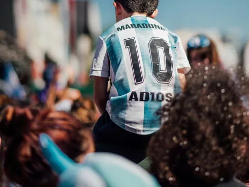 Enfant en maillot de foot argentin « Maradona 10 » sur les épaules d'un adulte, dans une foule ensoleillée.