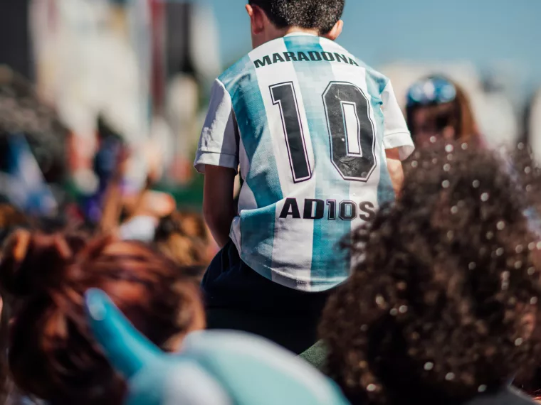 Enfant en maillot de foot argentin « Maradona 10 » sur les épaules d'un adulte, dans une foule ensoleillée.