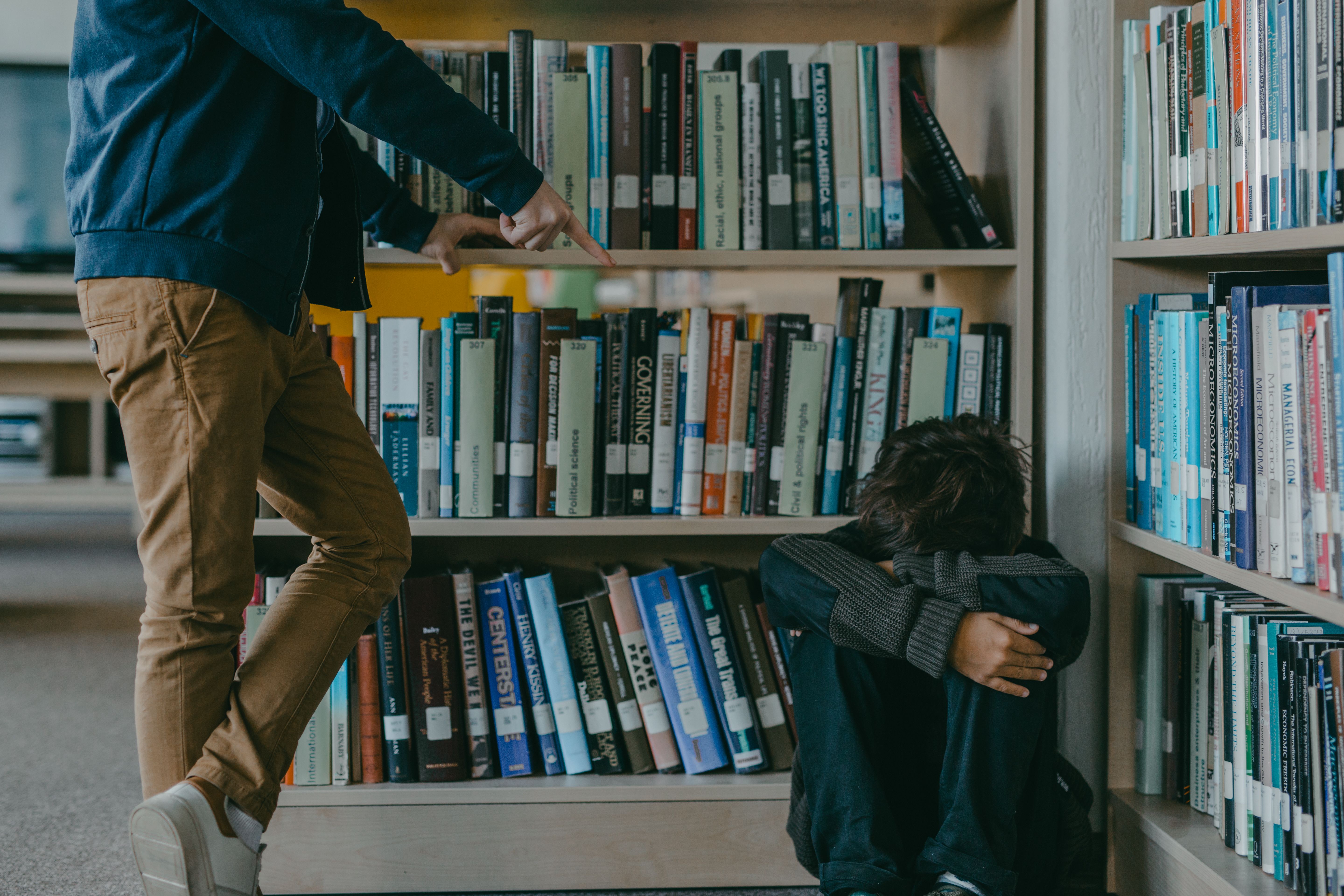 Harcèlement scolaire que faire en tant que parent : un enfant assis par terre dans une bibliothèque, la tête dans les bras, pendant qu’un autre élève le montre du doigt